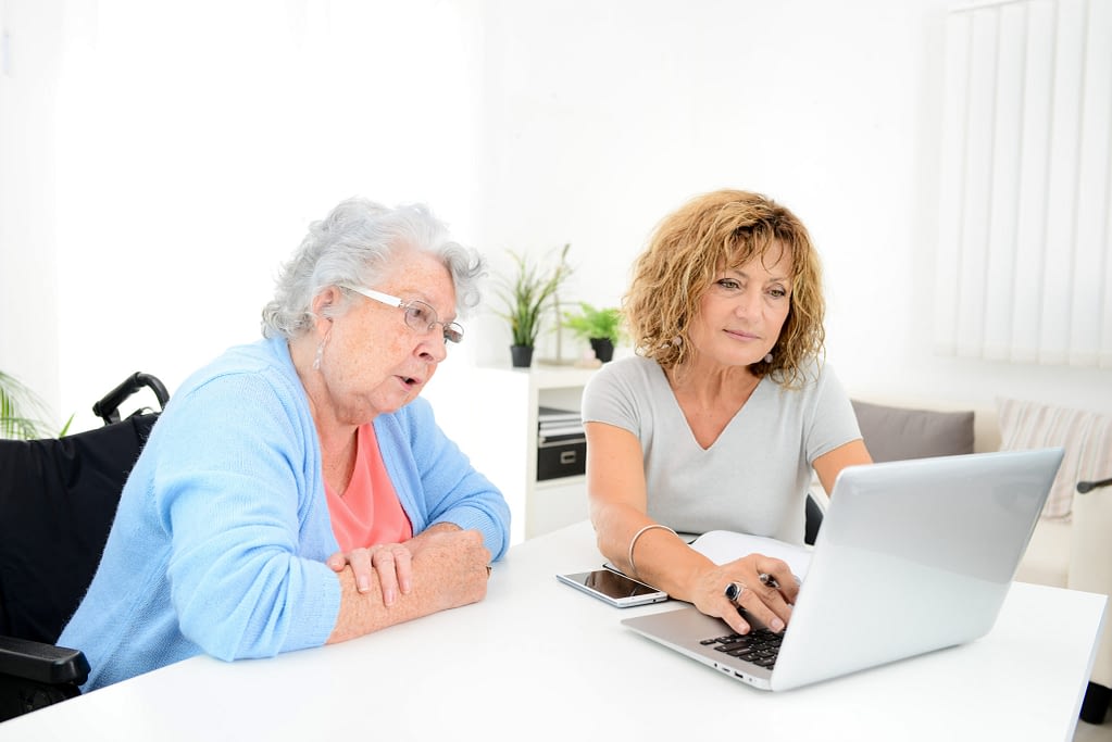 mature woman helping assisted elderly senior female with administrative procedures and paperwork on internet with a laptop computer at home